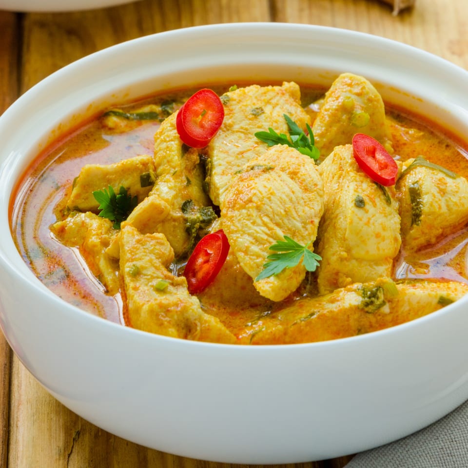 Chicken curry with red chili slices and parsley in a white bowl on a wooden table.