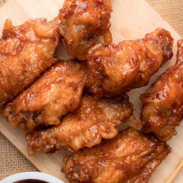 Close-up of several glistening, golden-brown chicken wings on a light wooden board, with a small bowl of sauce.