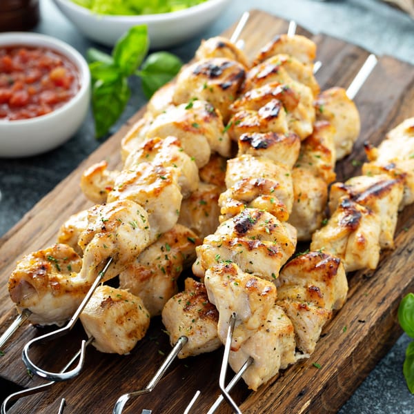 Grilled chicken kabobs on a wooden board, with salsa and guacamole in the background.